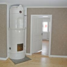 Biały, ceramiczny piec kaflowy w rogu pokoju z jasną podłogą i beżowymi ścianami. Widoczne otwarte, białe drzwi prowadzące do kolejnego pomieszczenia z oknem.