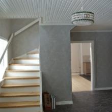 Klatka schodowa z drewnianymi schodami i szarą, teksturowaną ścianą. Widoczny fragment sufitu z boazerią, ozdobna lampa wisząca i drzwi do kolejnego pomieszczenia z jasną podłogą. Przy schodach...