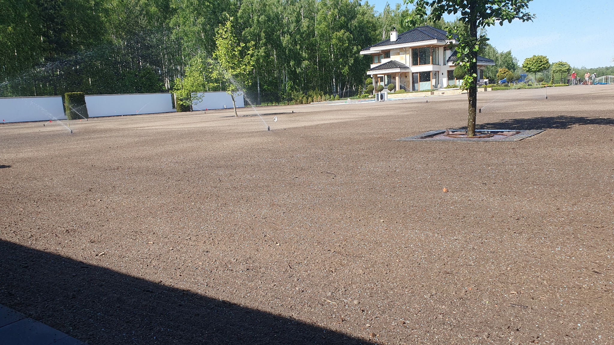 Rozległy teren po niwelacji, przygotowany pod trawnik, z systemem nawadniania w trakcie pracy. W tle nowoczesny dom i linia drzew.