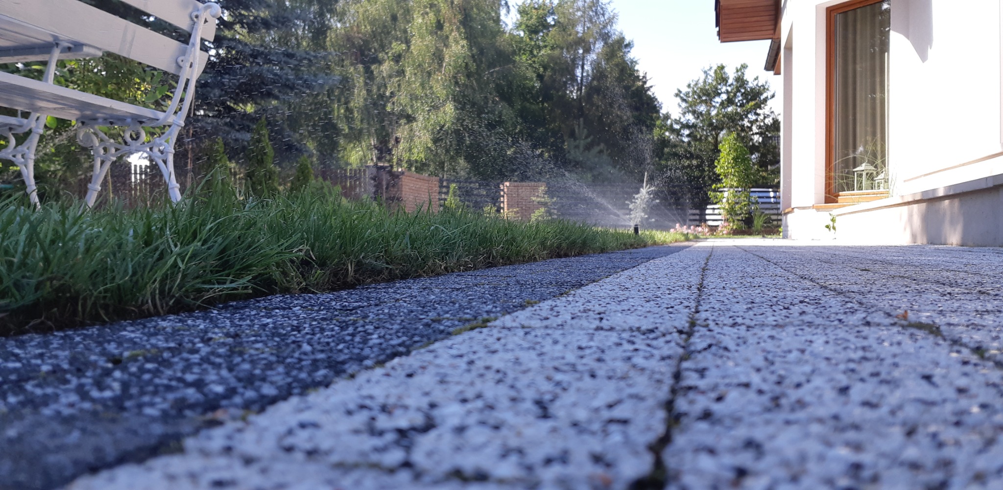 Niski kąt ujęcia kostki brukowej, trawnika z systemem nawadniania w ogrodzie. W tle biała ławka i budynek. Drobny deszcz zraszaczy.