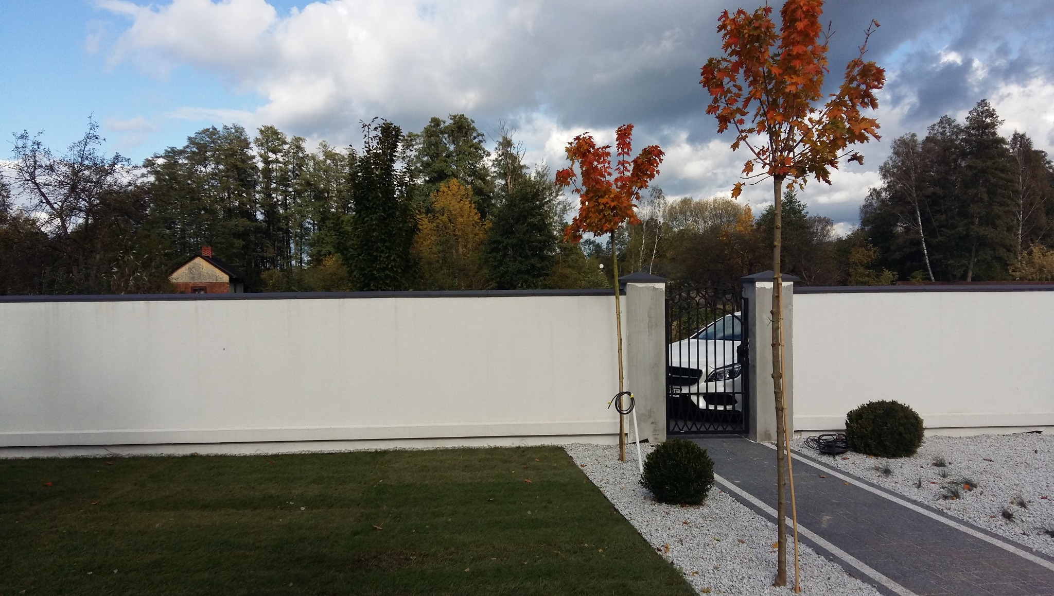 Nowoczesne ogrodzenie z bramą i furtką, obsadzone młodymi drzewkami o pomarańczowych liściach. Trawnik i ścieżka z jasnego żwiru. W tle drzewa i budynek.