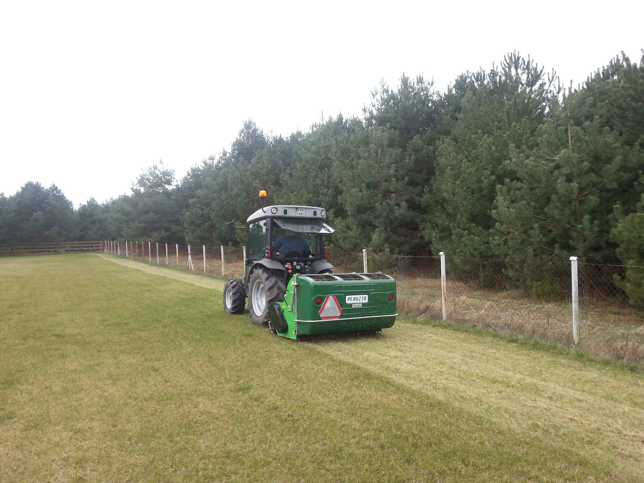 Zielony traktor Peruzzo z kosiarką rotacyjną na łące, świeżo skoszony pas trawy, widok z tyłu, drzewa w tle, ogrodzenie z siatki.