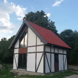 Nowoczesny dom z elementami muru pruskiego, czerwony dach, duże okno z odbiciem nieba, otoczony zielenią i drzewami.