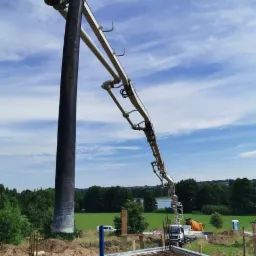 Wylewanie betonu na fundament domu z użyciem pompy do betonu; widoczna stalowa siatka zbrojeniowa, szalunki i krajobraz w tle.
