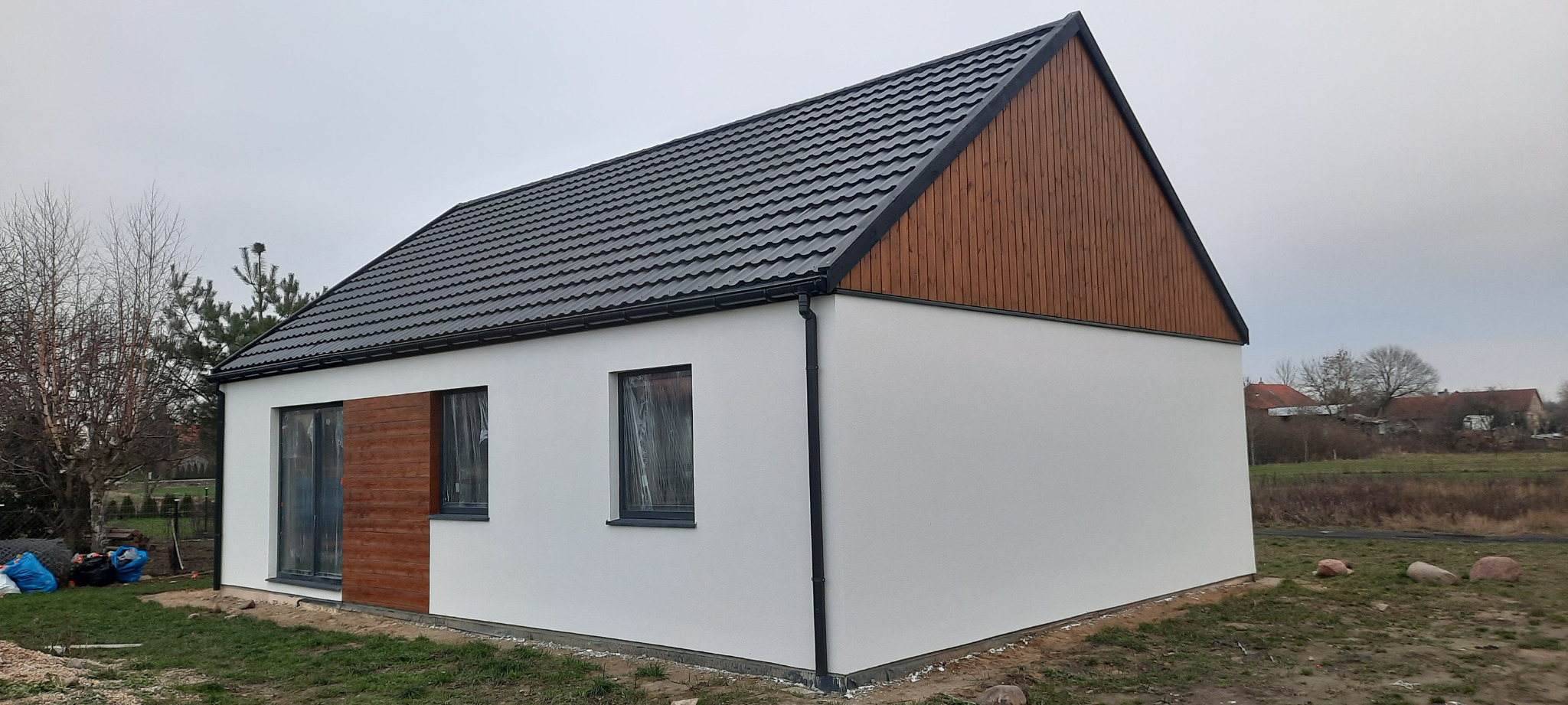 Gotowy, parterowy budynek z białymi ścianami i ciemnym dachem z blachodachówki, z drewnianym szczytem i oknami zabezpieczonymi folią, otoczony trawnikiem i zaroślami.