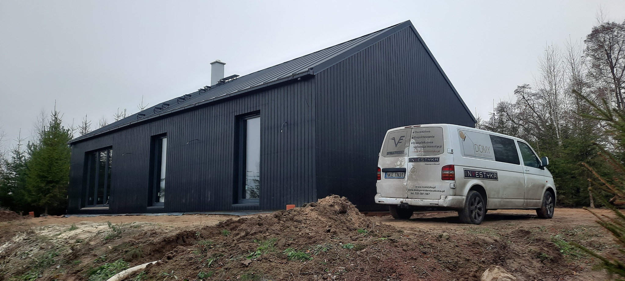 Nowoczesny, czarny dom z drewnianą elewacją i metalowym dachem, widziany z zewnątrz, obok stoi brudny biały bus firmy budowlanej z widocznym logo i numerem rejestracyjnym na tle drzew.