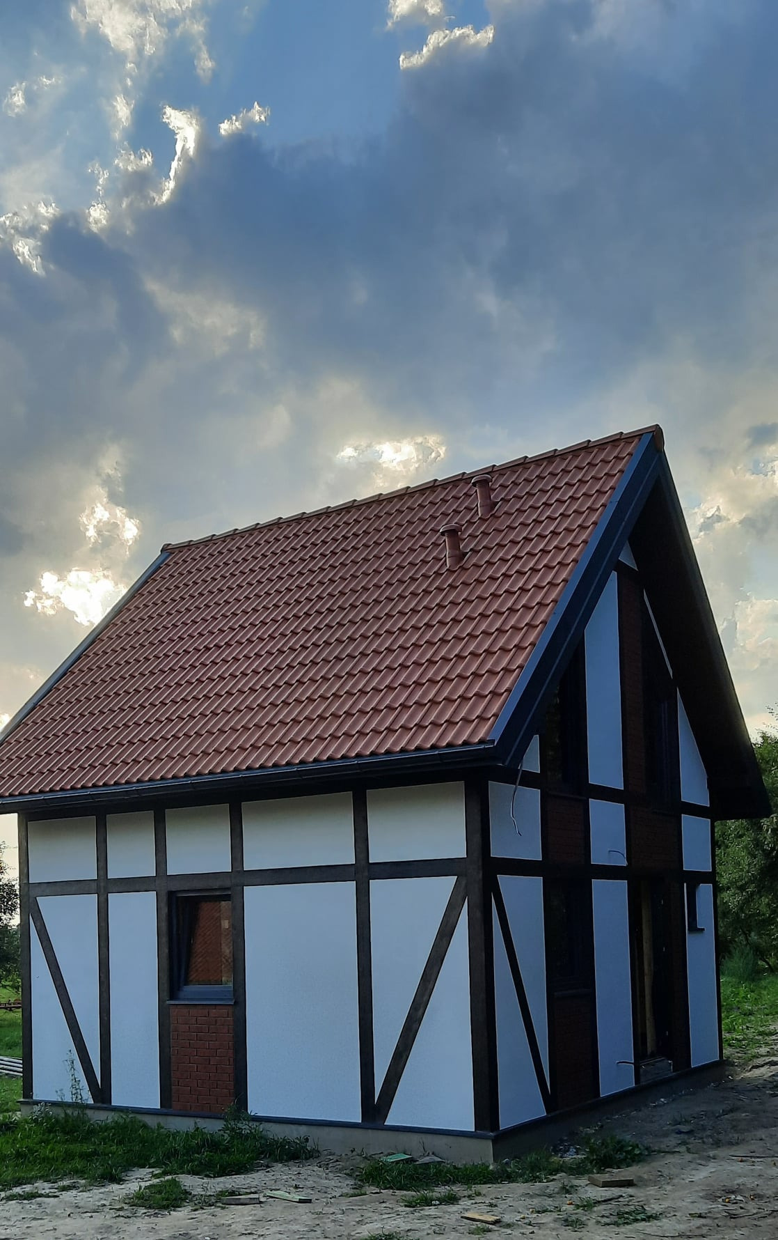 Niski, parterowy budynek w stylu szachulcowym z czerwoną dachówką, widoczny z perspektywy żabiej, na tle pochmurnego nieba z promieniami słońca przedzierającymi się przez chmury.