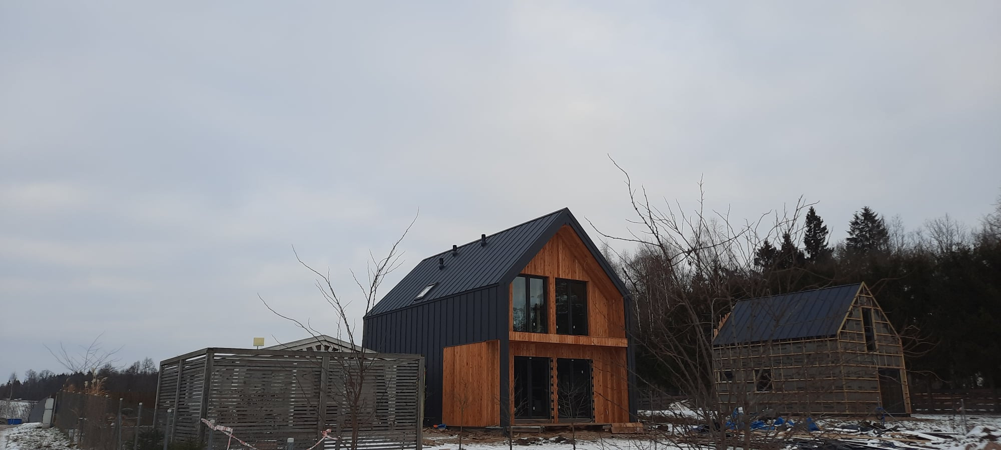 Nowoczesny dom z drewnianą elewacją i ciemnym metalowym dachem w trakcie budowy, widoczne rusztowania drugiego budynku w tle.