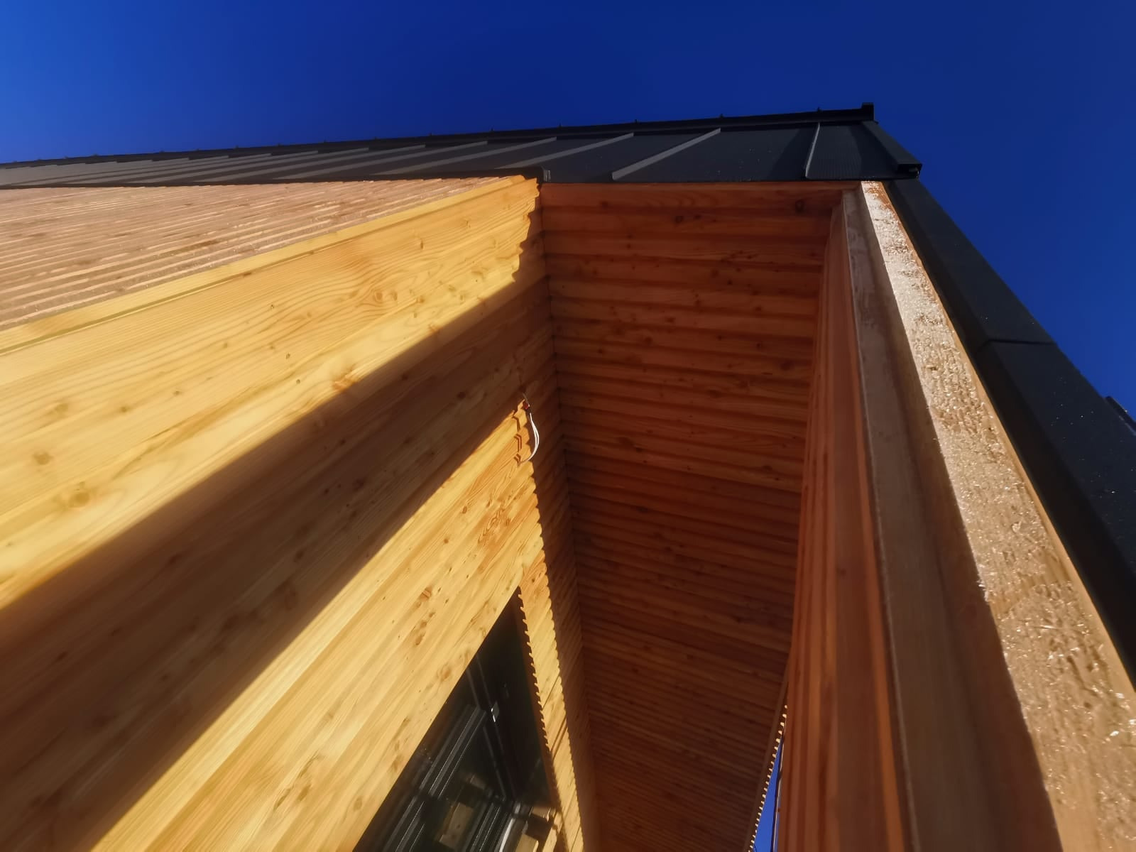 Surowa konstrukcja domu szkieletowego z widocznym drewnianym szkieletem, czarnym dachem i fragmentem okna, sfotografowana pod kątem w słoneczny dzień.