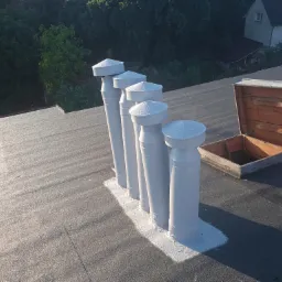 Czarny dach pokryty papą z pięcioma białymi kominkami wentylacyjnymi z kroplami wody na daszkach i otwartym włazem dachowym. W tle drzewa i dom.