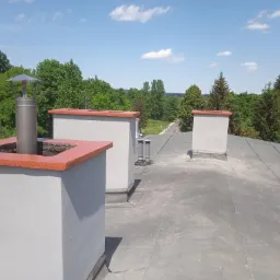 Płaski dach z widocznymi kominami wentylacyjnymi i narzędziami dekarskimi, otoczony zielenią drzew i błękitnym niebem.