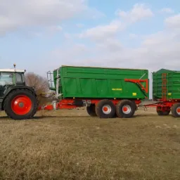 Zielony traktor Fendt z dwiema przyczepami Pronar na polu, błękitne niebo z chmurami w tle.