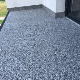 Szara, kamienista powierzchnia balkonu z widocznym odpływem, obrzeżem i fragmentem okna balkonowego oraz zielonym otoczeniem w tle.