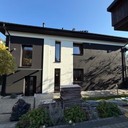 Termodocieplenia.pl - Dwukondygnacyjny dom z elewacją w kolorze białym i antracytowym, z widocznymi materiałami budowlanymi i workiem na śmieci przed budynkiem, w słoneczny dzień.