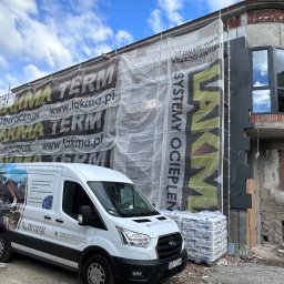 Termodocieplenia.pl - Budynek w trakcie prac elewacyjnych: ocieplanie styropianem grafitowym, widoczne rusztowania z banerem reklamowym firmy, zaparkowany firmowy bus z reklamą usług, palety z workami zaprawy klejowej