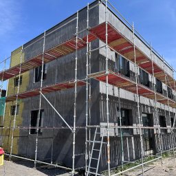 Termodocieplenia.pl - Dwupiętrowy budynek w trakcie prac elewacyjnych, z widocznym rusztowaniem, częściowo ocieploną ścianą w kolorze żółtym i szarym, oraz paczką wełny mineralnej u podstawy.