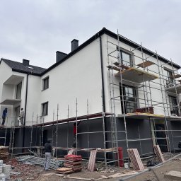 Termodocieplenia.pl - Dwupiętrowy dom w trakcie prac elewacyjnych: białe ściany na górze, szare na dole, otoczony rusztowaniami, na których pracują robotnicy. Na pierwszym planie porozrzucane materiały budowlane.