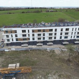 Termodocieplenia.pl - Wielopiętrowy budynek z panelami słonecznymi na dachu, widziany z lotu ptaka. Widoczne rusztowania i teren budowy. W tle zielone pola i lasy.