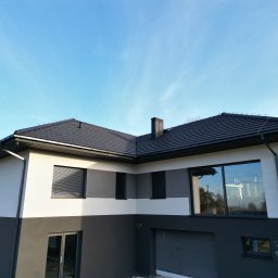 Termodocieplenia.pl - Dwukolorowa elewacja domu z grafitową dachówką i oknami z roletami zewnętrznymi. Jasne tło nieba.