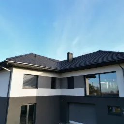 Dwukolorowa elewacja domu z grafitową dachówką i oknami z roletami zewnętrznymi. Jasne tło nieba.