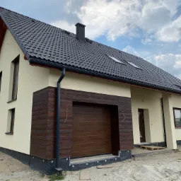 Nowoczesny dom jednorodzinny z beżową elewacją, brązowymi akcentami imitującymi drewno przy garażu i oknach, oraz ciemnoszarą dachówką, widoczny fragment fundamentów z izolacją.