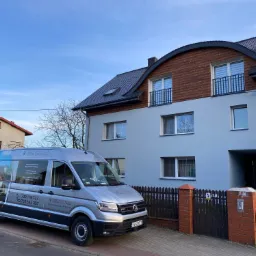 Ocieplony dom jednorodzinny z szarą elewacją i drewnianą górną częścią, na pierwszym planie firmowy bus z reklamą usług ocieplania elewacji.