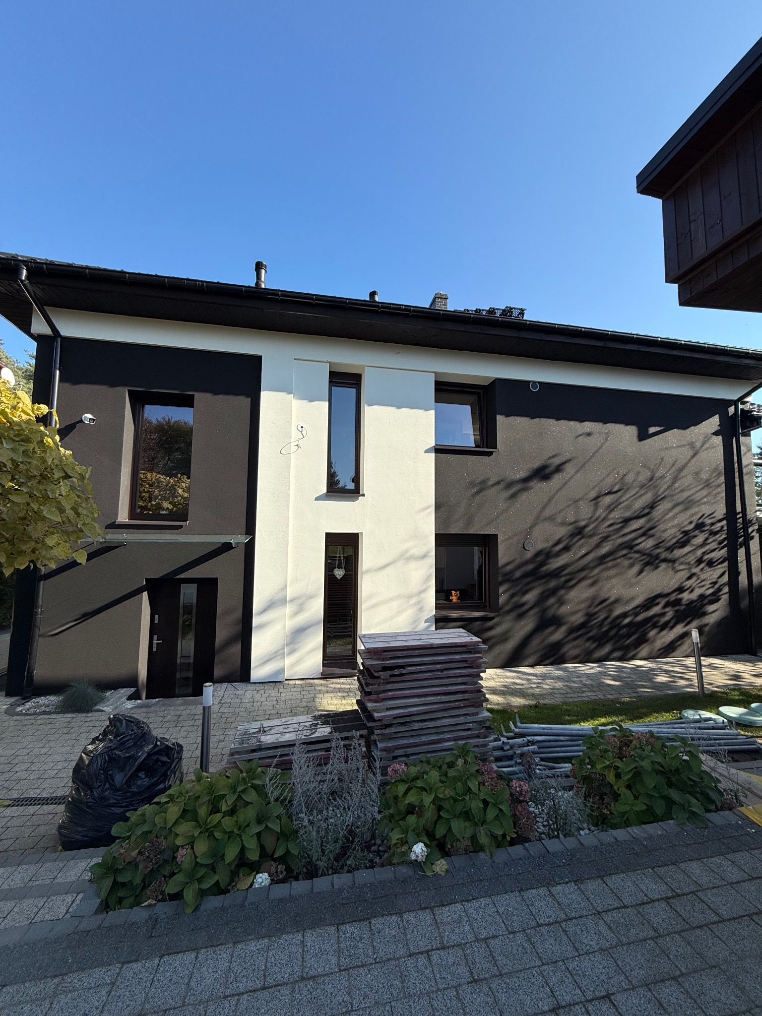 Dwukondygnacyjny dom z elewacją w kolorze białym i antracytowym, z widocznymi materiałami budowlanymi i workiem na śmieci przed budynkiem, w słoneczny dzień.