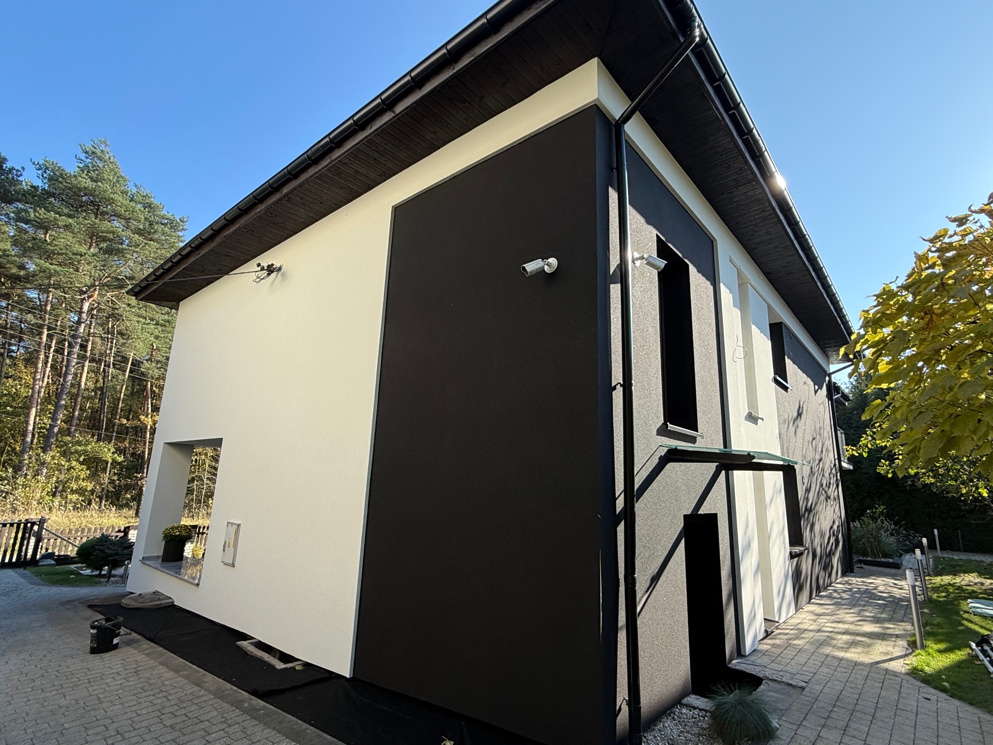 Nowoczesny dom jednorodzinny z elewacją w kolorze czerni i bieli, wyposażony w system monitoringu wizyjnego, otoczony zielenią i brukowanym podjazdem.