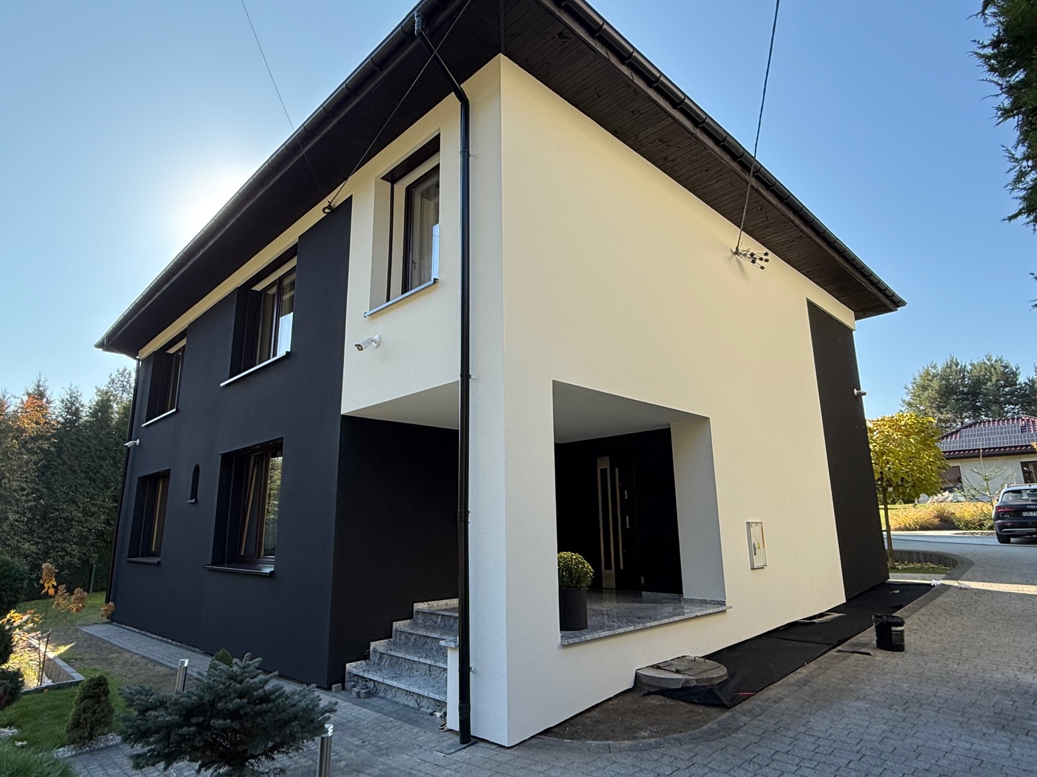 Dwukolorowy dom z elewacją w tonacji bieli i grafitu, widok z zewnątrz w słoneczny dzień, z widocznymi schodami wejściowymi z jasnego kamienia i zadbanym otoczeniem.