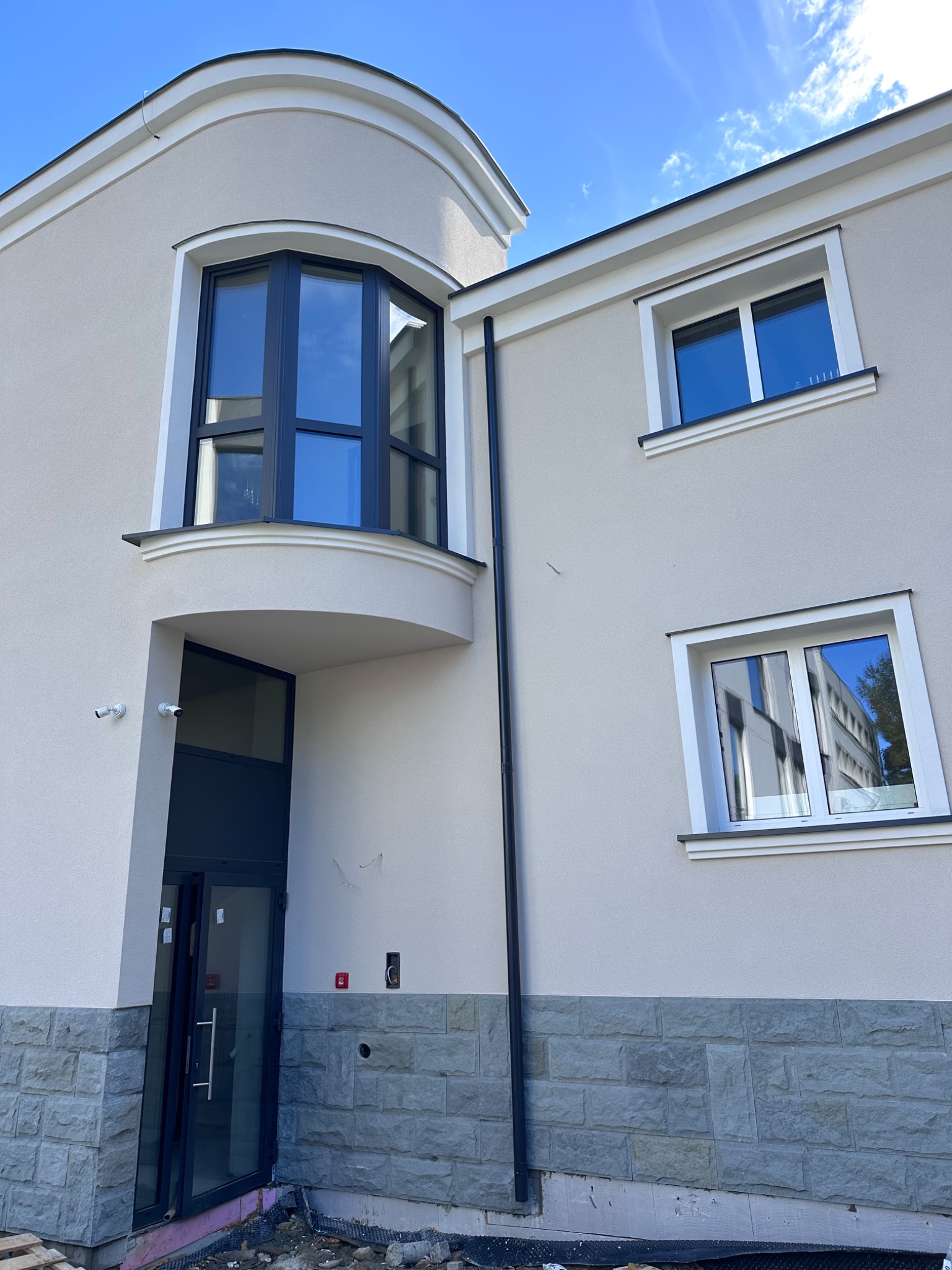 Nowoczesny budynek z jasną elewacją, ozdobnym wykuszem okiennym i kamienną podmurówką, widoczne dwa okna i drzwi wejściowe, podgląd na niezagospodarowany teren wokół budynku.