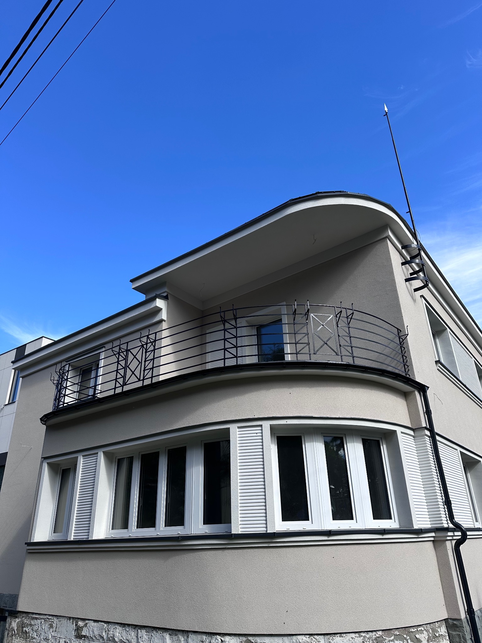 Elewacja budynku w stylu modernistycznym z zaokrąglonym balkonem i metalową balustradą, widok z dołu na tle błękitnego nieba.