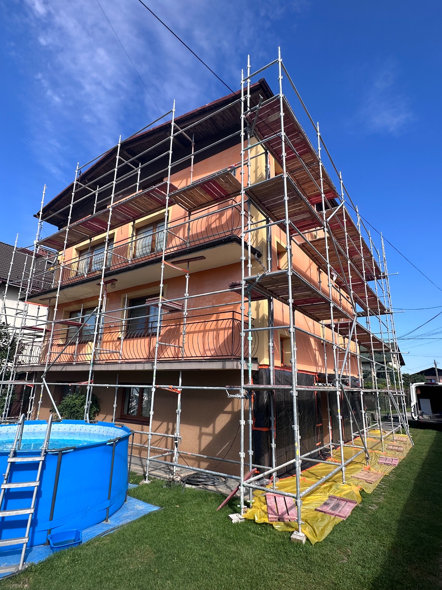 Dom jednorodzinny w trakcie prac elewacyjnych, otoczony rusztowaniem, z widocznym basenem ogrodowym w pobliżu.