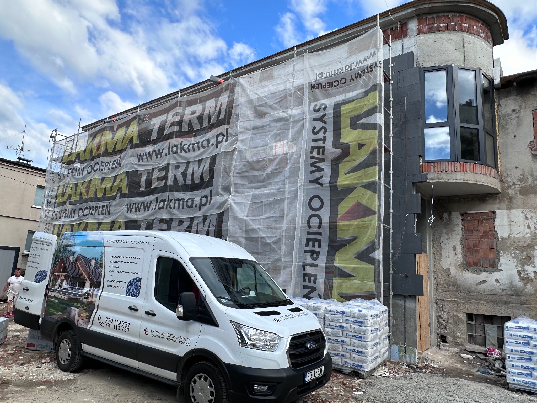 Budynek w trakcie prac elewacyjnych: ocieplanie styropianem grafitowym, widoczne rusztowania z banerem reklamowym firmy, zaparkowany firmowy bus z reklamą usług, palety z workami zaprawy klejowej