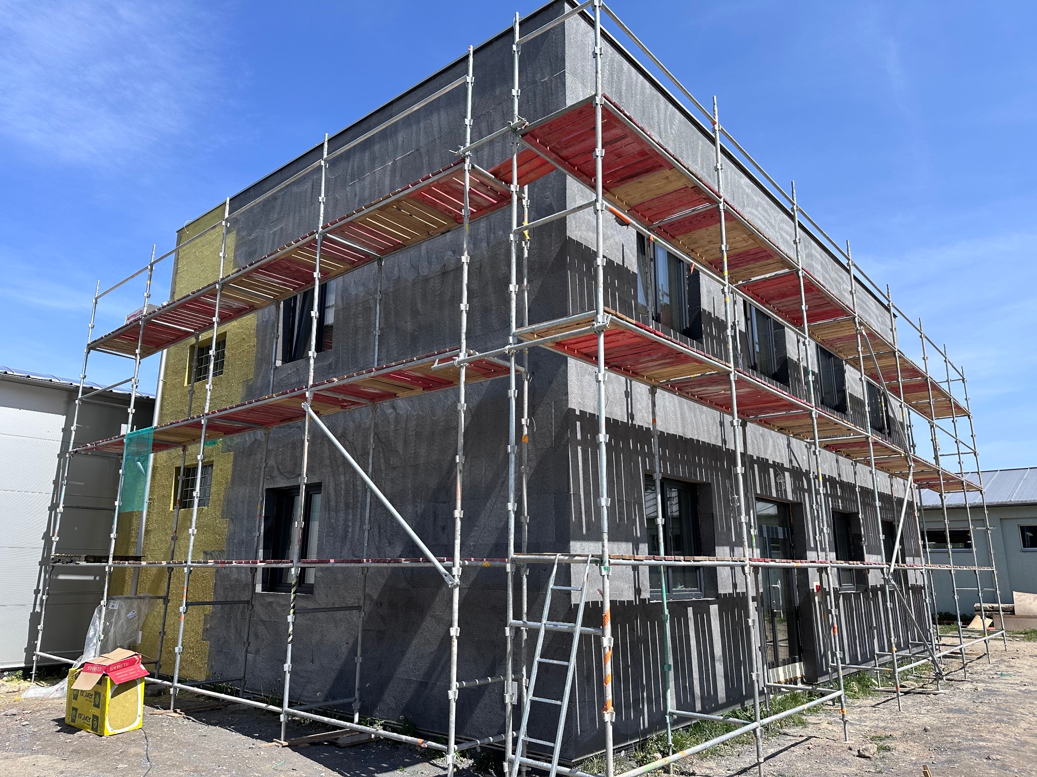 Dwupiętrowy budynek w trakcie prac elewacyjnych, z widocznym rusztowaniem, częściowo ocieploną ścianą w kolorze żółtym i szarym, oraz paczką wełny mineralnej u podstawy.