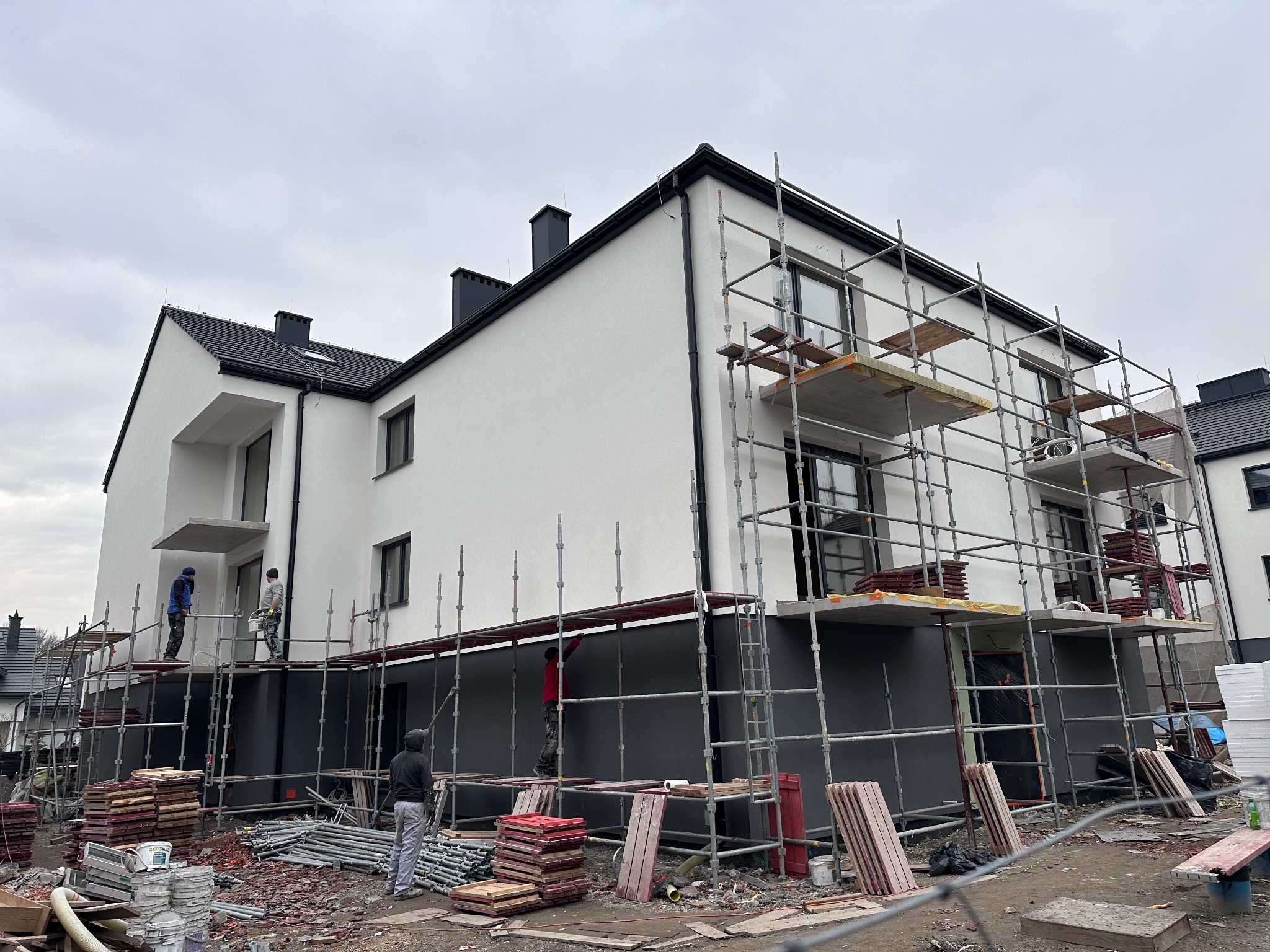 Dwupiętrowy dom w trakcie prac elewacyjnych: białe ściany na górze, szare na dole, otoczony rusztowaniami, na których pracują robotnicy. Na pierwszym planie porozrzucane materiały budowlane.