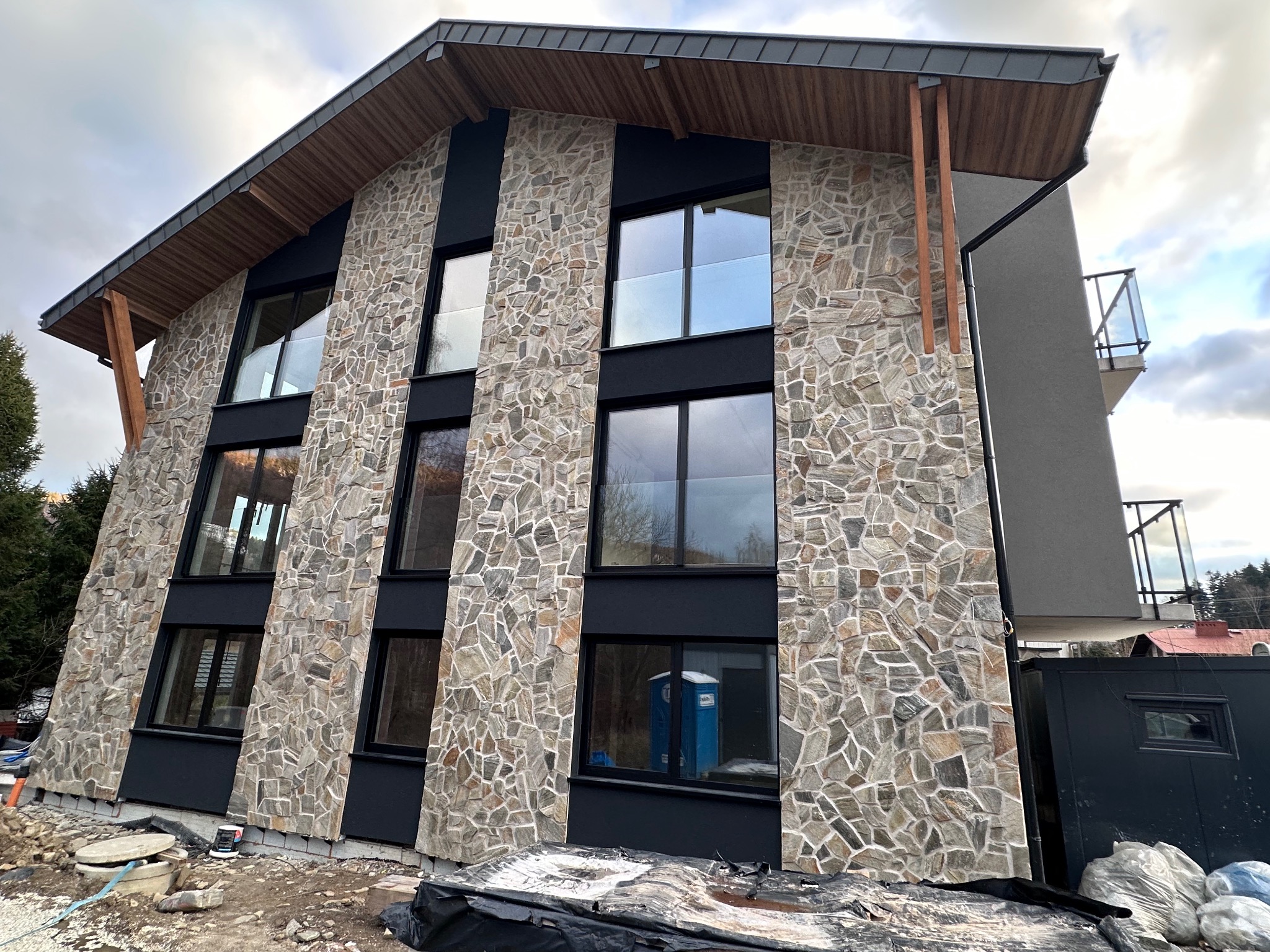 Dwupiętrowy dom z elewacją z kamienia naturalnego w odcieniach szarości i beżu, z czarnymi ramami okiennymi, drewnianym zadaszeniem i widoczną toaletą przenośną w odbiciu okna na placu budowy.