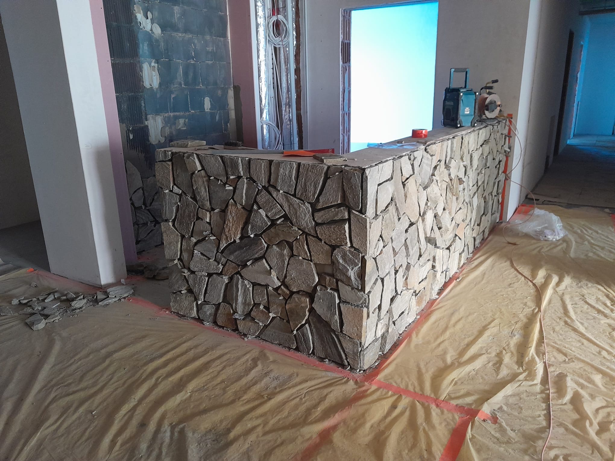 Wnętrze w trakcie wykańczania: fragment ściany wyłożony nieregularnym kamieniem, folia ochronna na podłodze, widoczne instalacje i narzędzia na blacie.