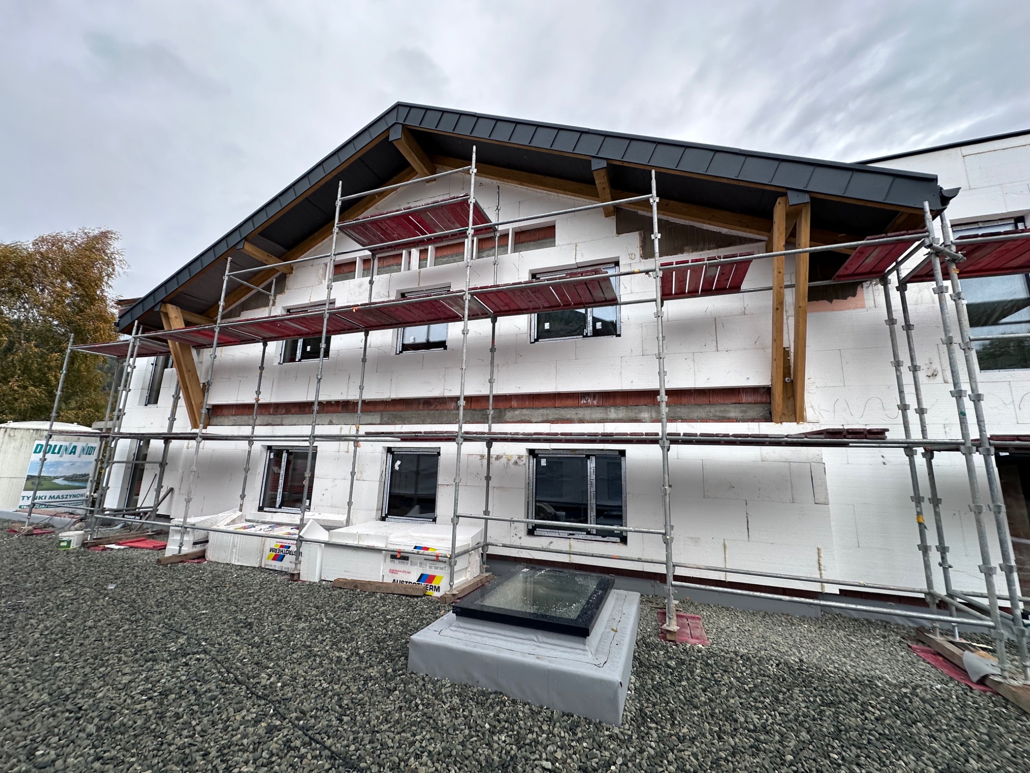 Dom w trakcie prac z ociepleniem elewacji, widoczne rusztowania, płyty styropianowe, okna i przygotowane podłoże z kamyczków.