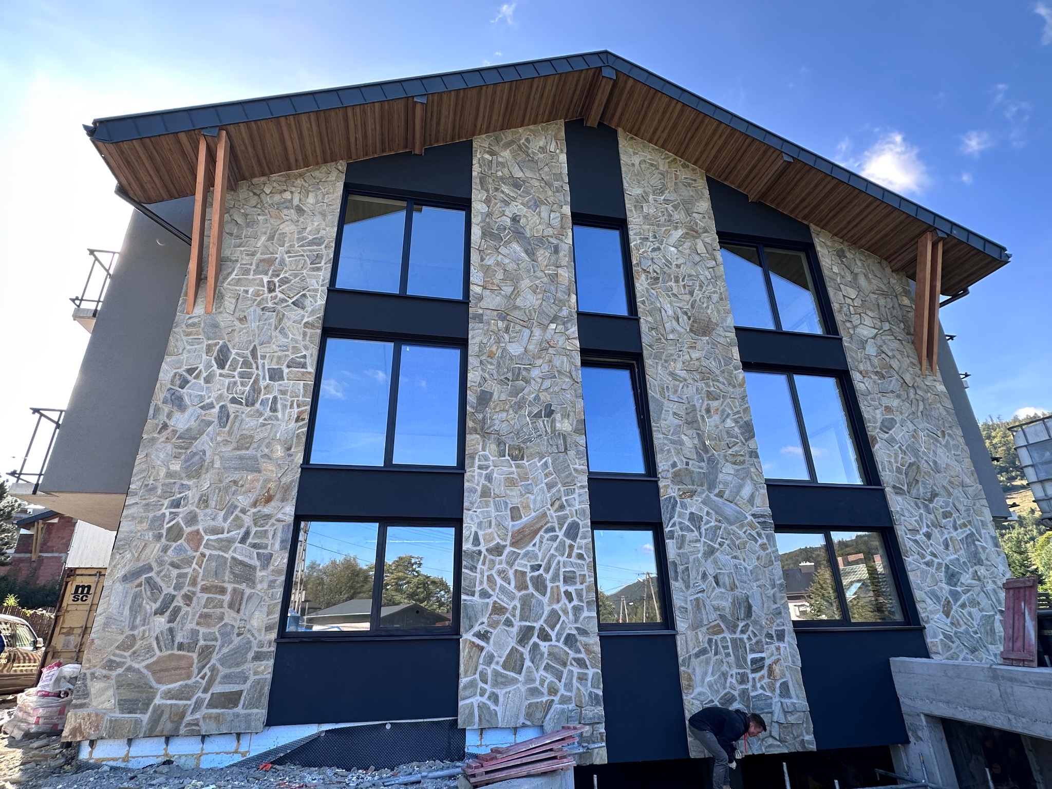 Nowoczesny budynek z elewacją z kamienia naturalnego i czarnymi panelami, widoczne okna odbijające niebo, w tle zieleń, prace wykończeniowe w toku.