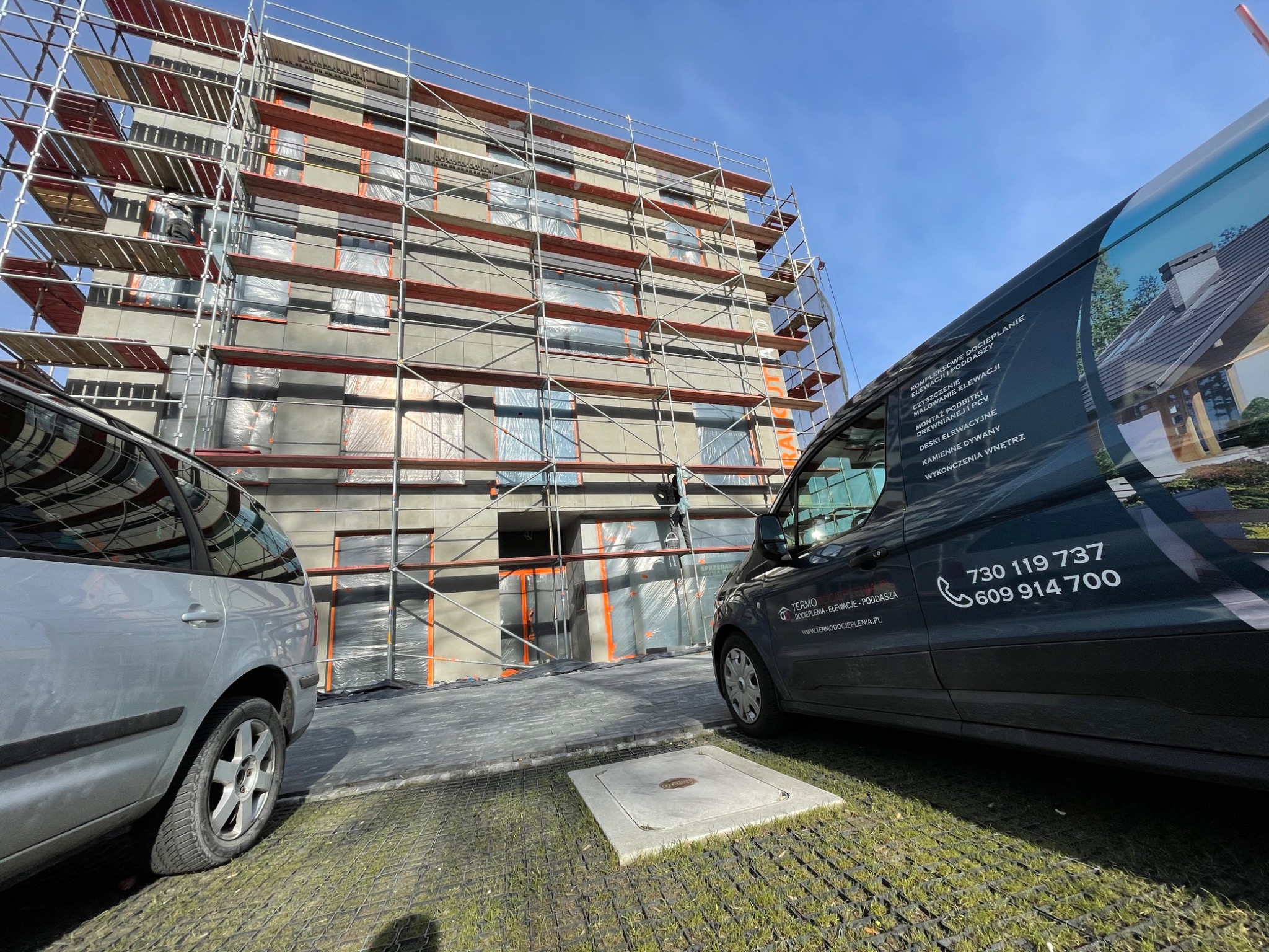 Budynek wielorodzinny w trakcie prac elewacyjnych, rusztowania pokrywają fasadę, okna zabezpieczone folią, obok zaparkowany samochód osobowy i firmowy bus z reklamą usług termoizolacyjnych.