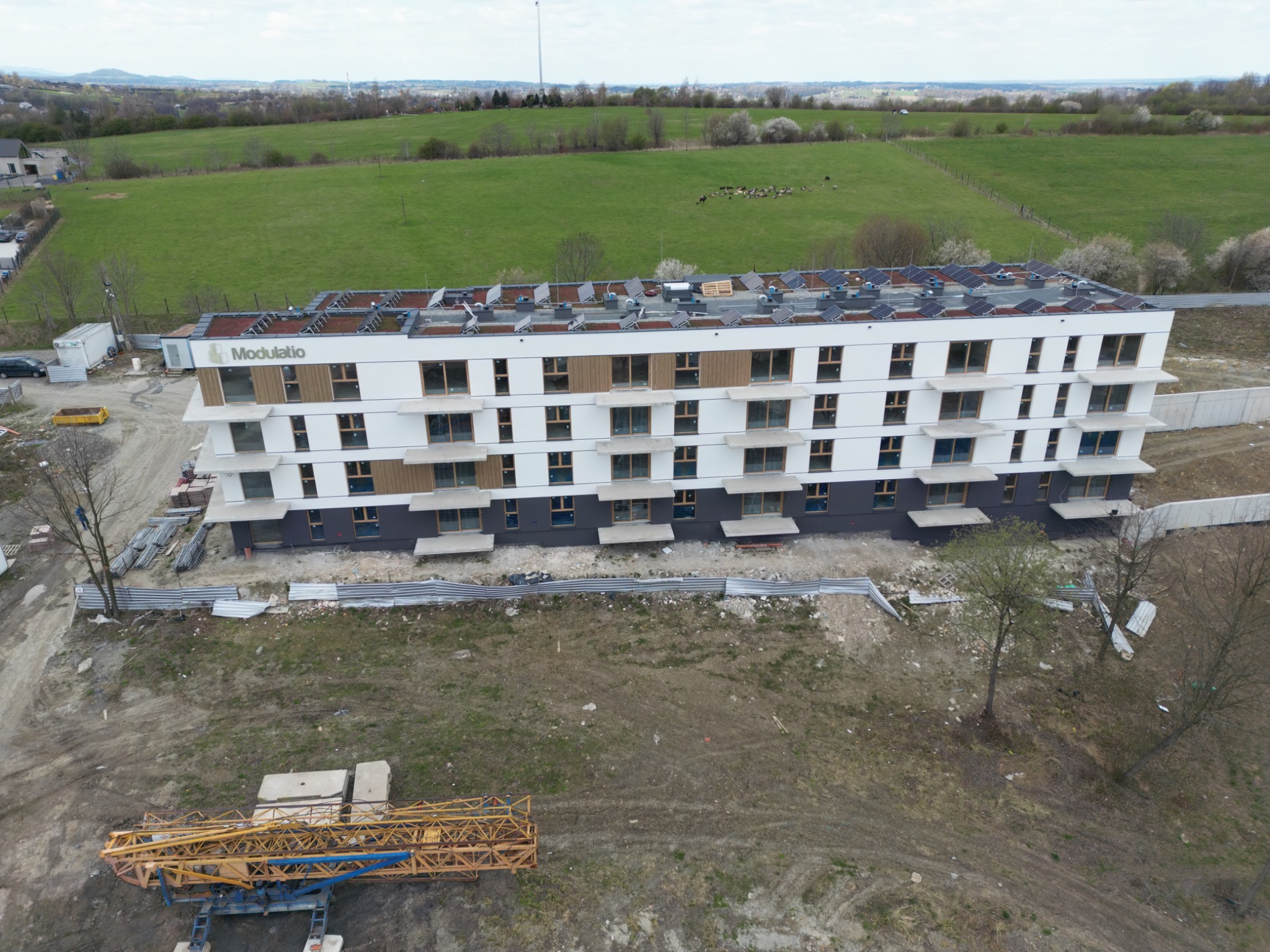 Wielopiętrowy budynek z panelami słonecznymi na dachu, widziany z lotu ptaka. Widoczne rusztowania i teren budowy. W tle zielone pola i lasy.