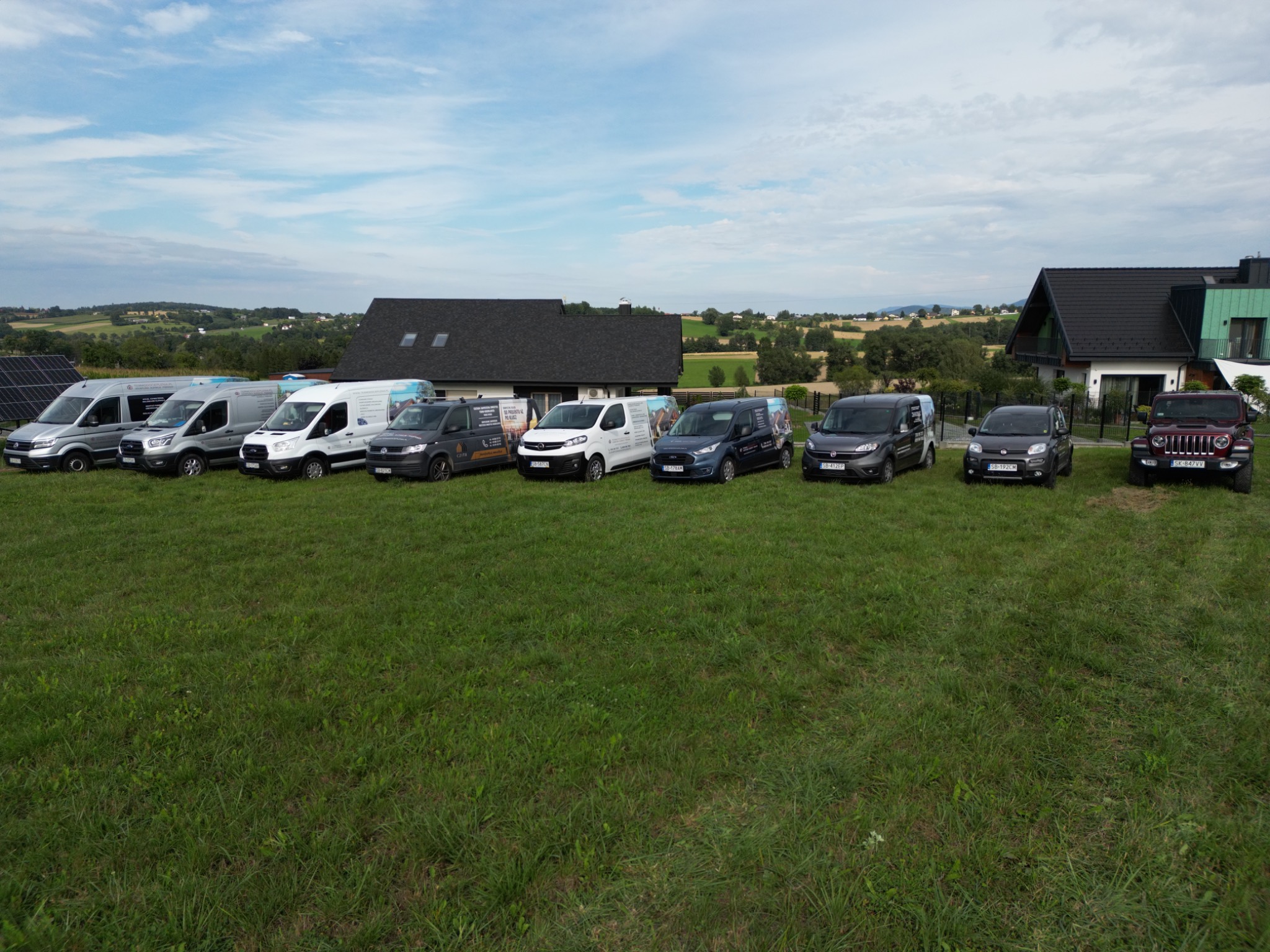 Flota firmowych samochodów na tle domu z panelami słonecznymi i malowniczym krajobrazem. Różne modele i kolory aut, każdy z logo firmy.