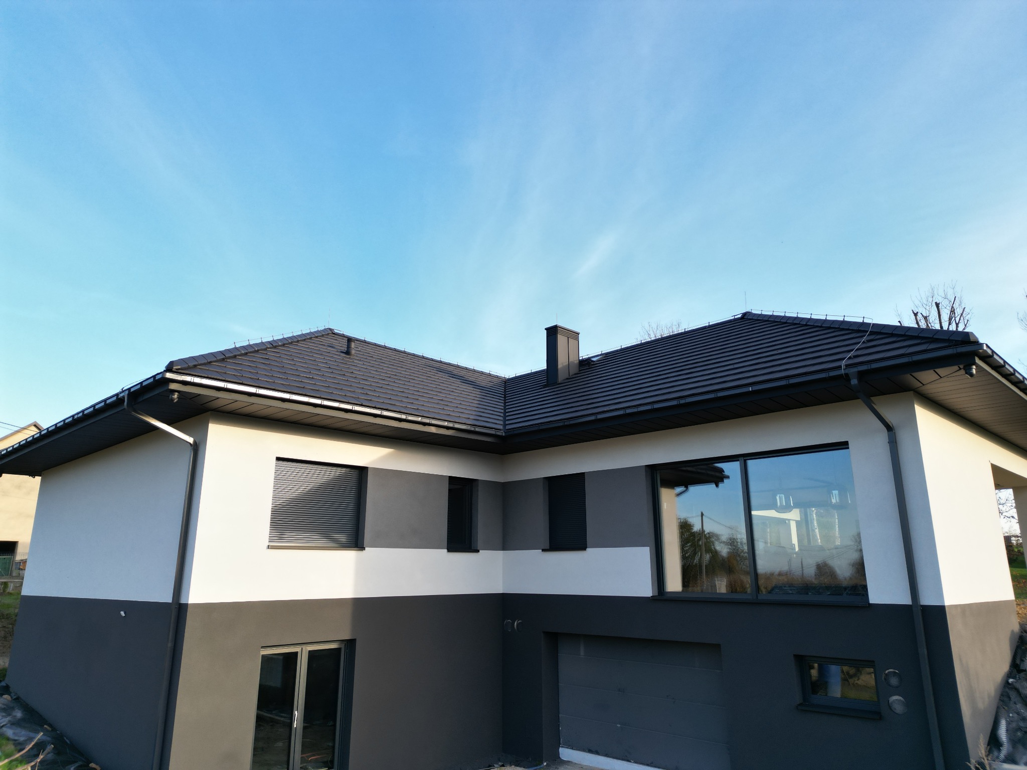 Dwukolorowa elewacja domu z grafitową dachówką i oknami z roletami zewnętrznymi. Jasne tło nieba.