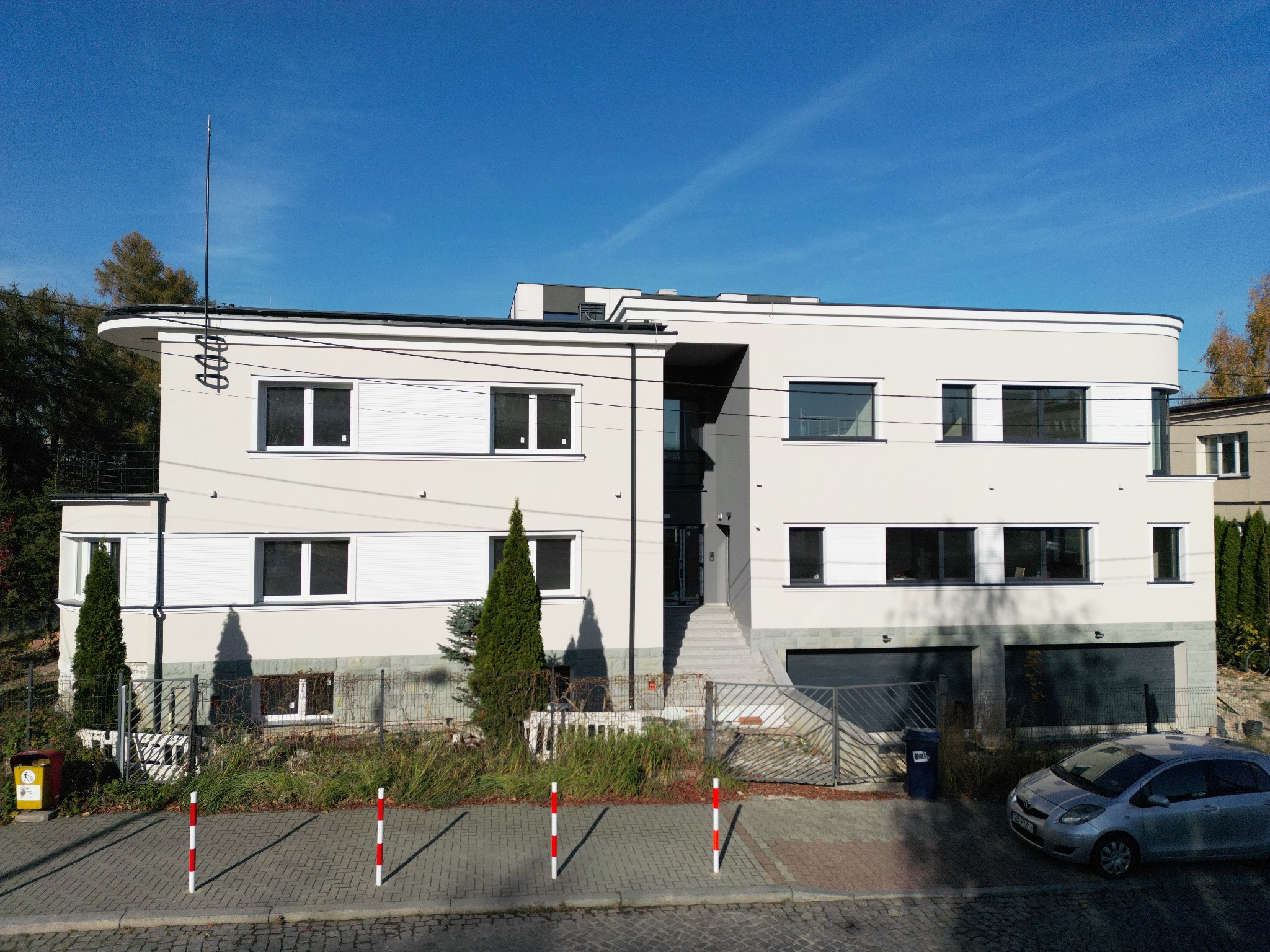 Dwupiętrowy dom z jasną elewacją w stylu modernistycznym, z płaskim dachem, dużymi oknami i garażem. Na dachu widoczna antena. W tle błękitne niebo.