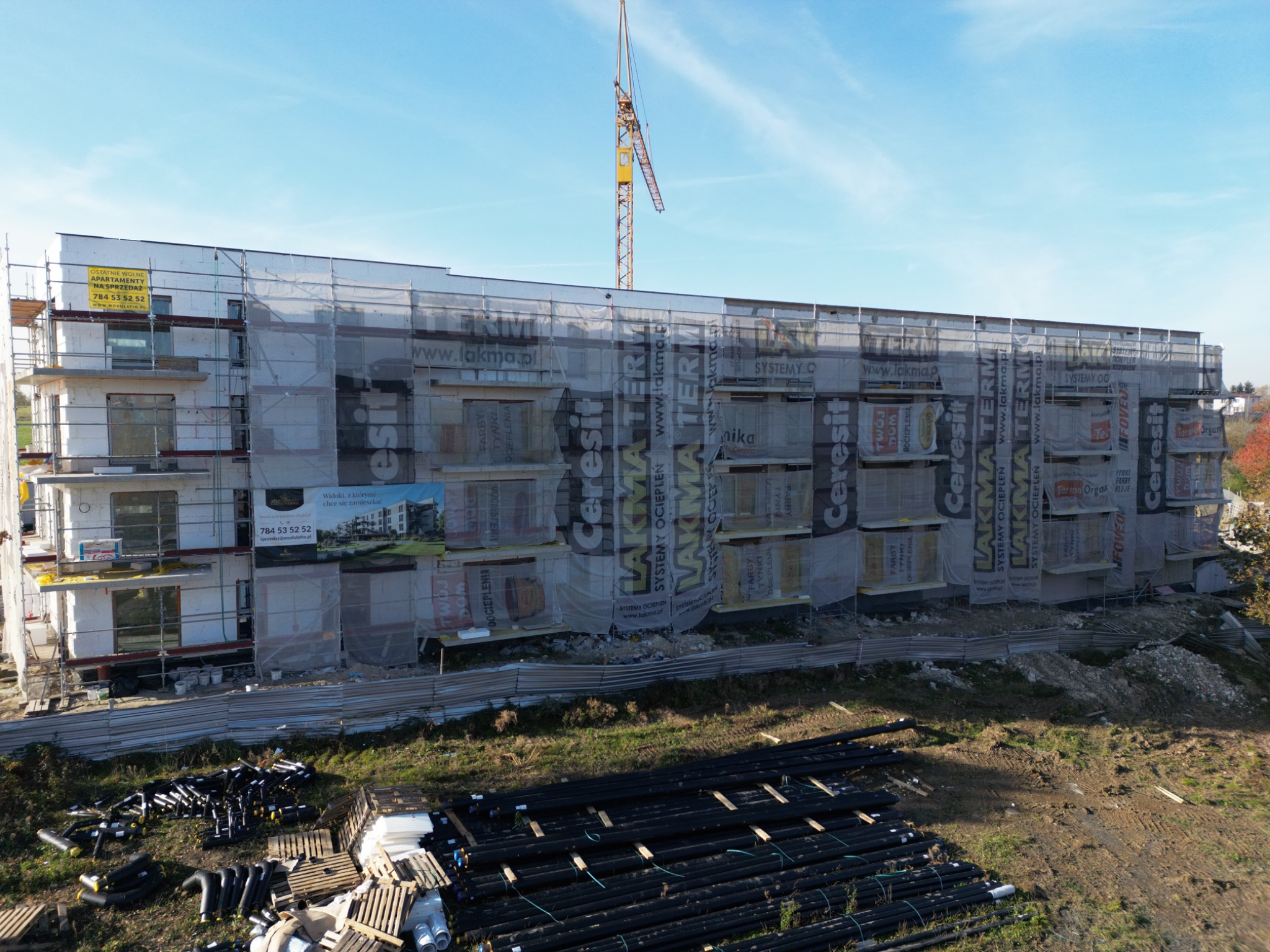 Wielopiętrowy budynek w trakcie ocieplania elewacji, osłonięty siatką z reklamami, widoczny dźwig budowlany i materiały budowlane na ziemi.