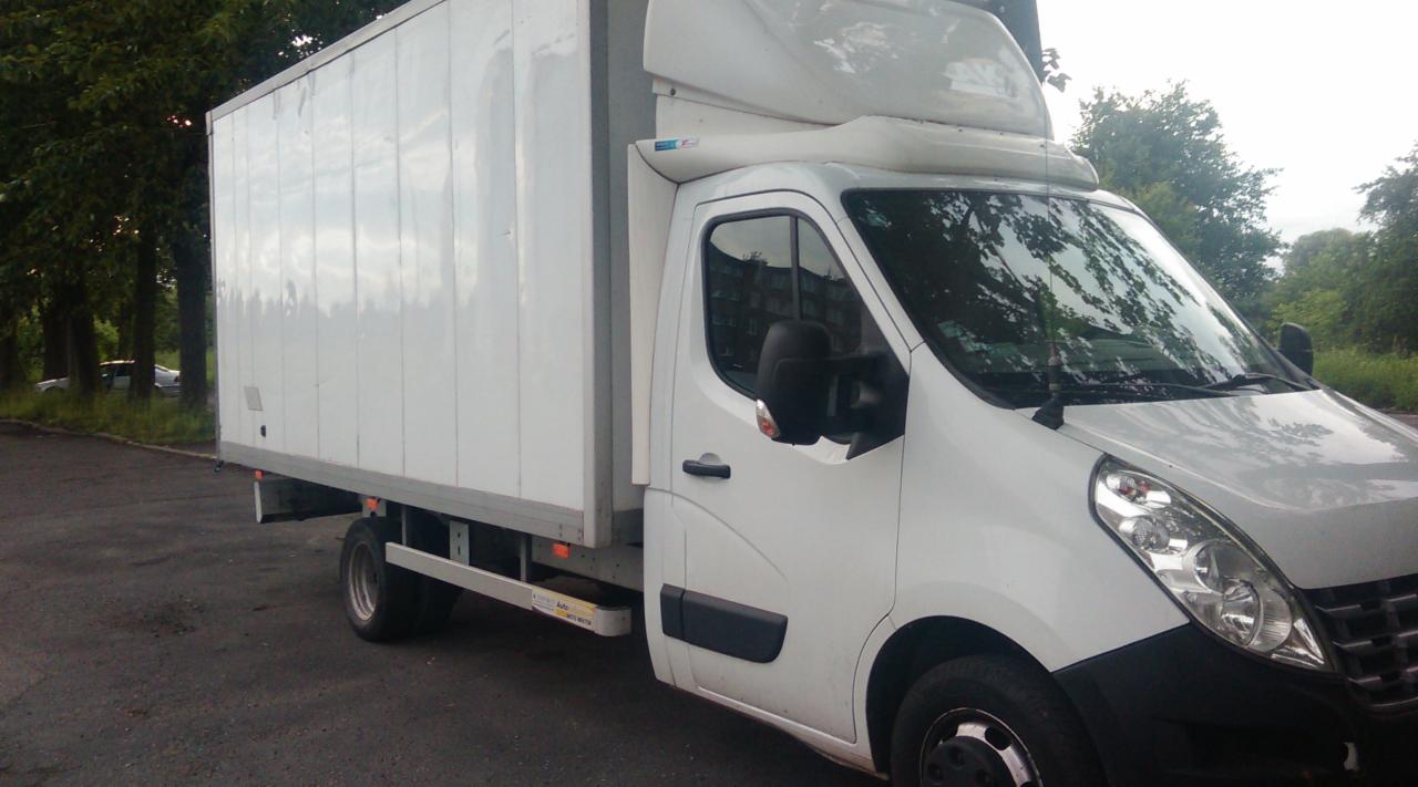 Biały samochód dostawczy z wysoką zabudową kontenerową, zaparkowany na asfaltowym placu w pobliżu drzew.