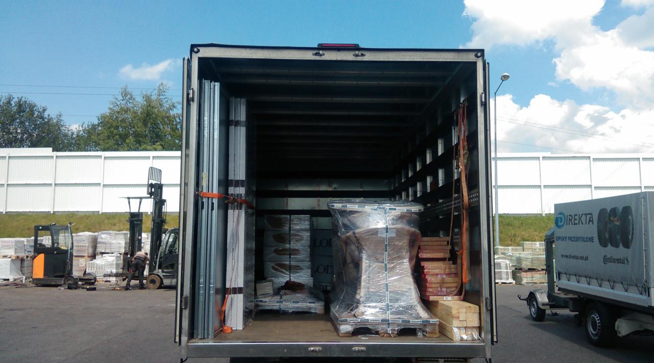 Wnętrze skrzyni ładunkowej wypełnione materiałami budowlanymi, zabezpieczonymi folią stretch, z widocznymi wózkiem widłowym i pracownikiem na zewnątrz, na tle białego ekranu akustycznego.