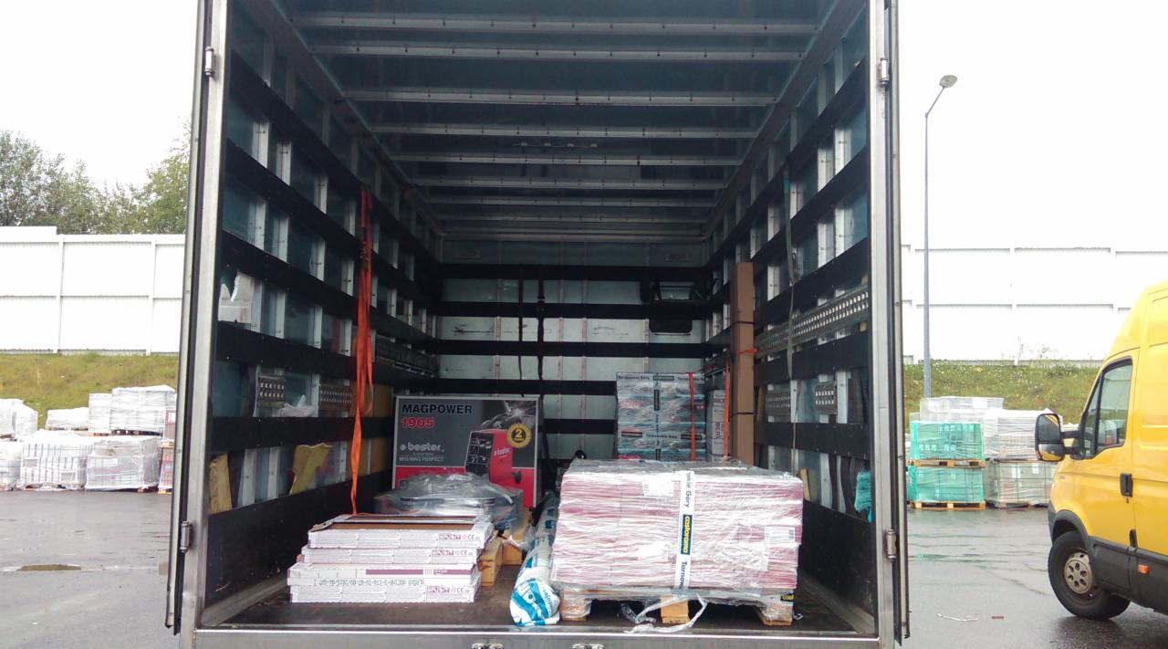 Wnętrze paki busa załadowane paletami z materiałami budowlanymi i urządzeniem Magpower 1905, widoczny fragment żółtego samochodu dostawczego na zewnątrz.