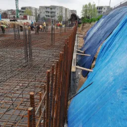 Siatka zbrojeniowa fundamentu z pionowymi prętami, widoczni pracownicy w oddali, zabezpieczenie wykopu plandeką.