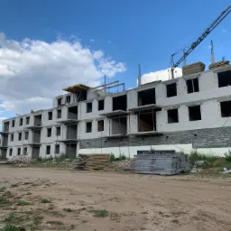 Wielopiętrowy budynek w stanie surowym, murowany z bloczków, widoczny dźwig budowlany w tle, przygotowane szalunki na ziemi.